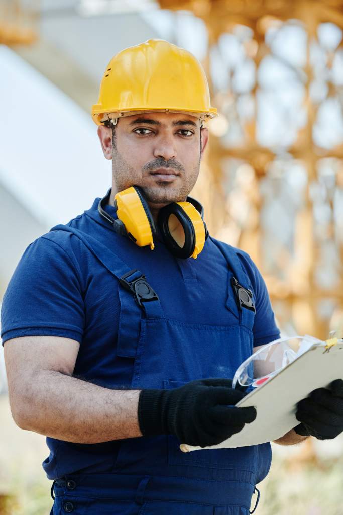 Das Bild zeigt einen Arbeiter auf einer Baustelle mit Schutzausrüstung. Er hat eine Checkliste in der Hand.