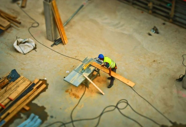 Das Bild zeigt, wie ein Arbeiter auf einer Baustelle alleine ein Brett schneidet. Das Bild ist aus der Vogelperspektive.